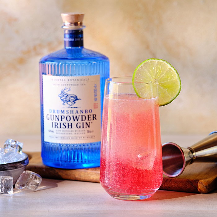 A blue bottle of Drumshanbo Gunpowder Irish Gin beside a pink cocktail garnished with a lime slice on a wooden board with ice cubes and a jigger.