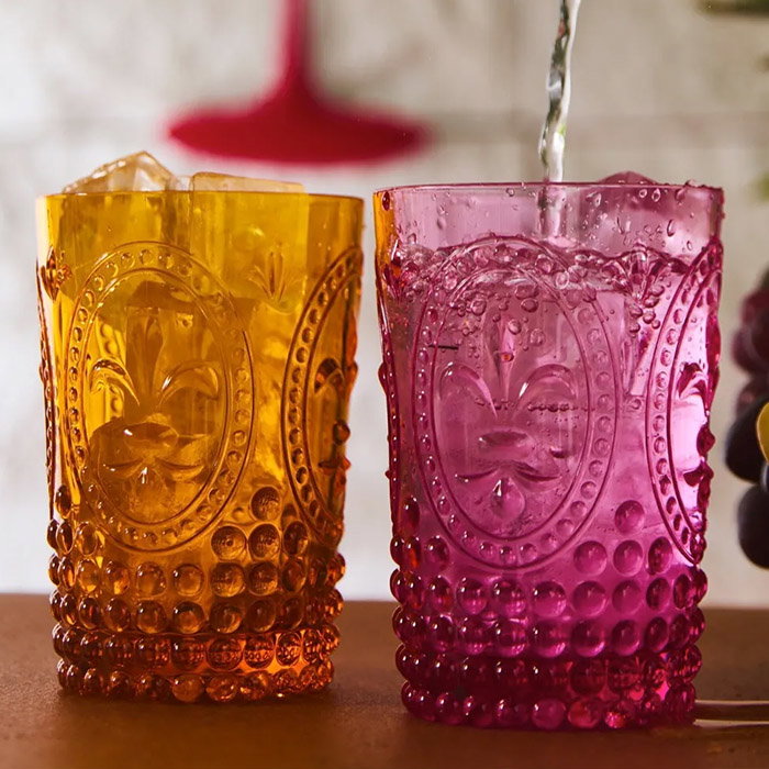 Two ornate vintage-style glasses, one amber with ice and one pink being filled with water, showcasing textured patterns and a fleur-de-lis design.