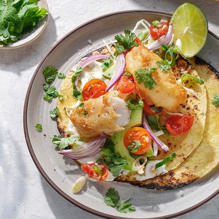 Grilled fish taco with cherry tomatoes, avocado, red onion, cilantro, cream sauce, and lime on a charred tortilla on a beige plate.