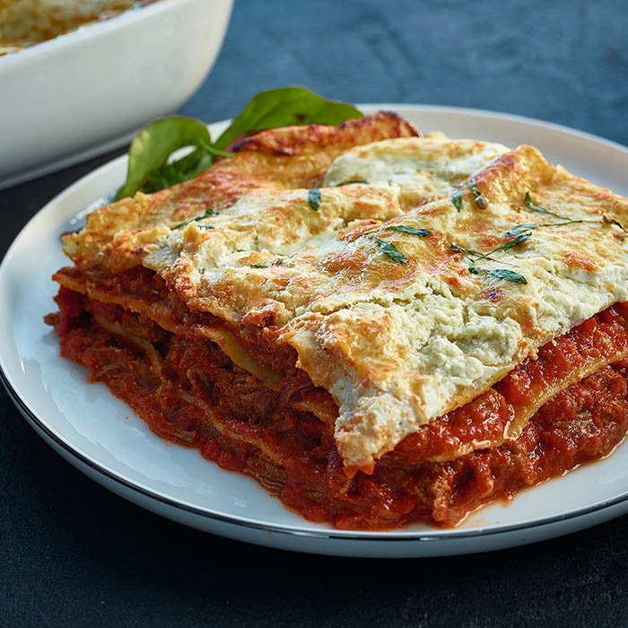 Close-up of a slice of layered lasagna with tomato sauce, ricotta cheese, and herbs on a white plate.