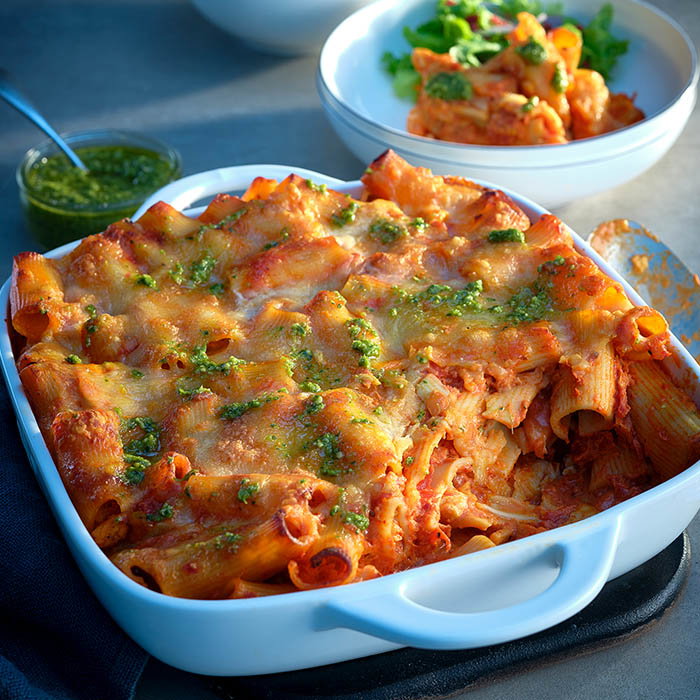 Baked rigatoni pasta in a white dish topped with melted cheese and green pesto, with a bowl of pasta salad and green sauce in the background.