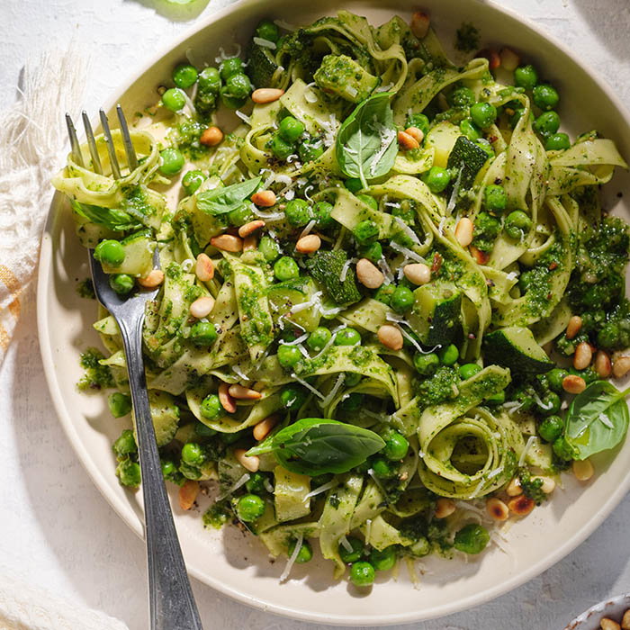 Plate of green pesto pasta with peas, zucchini, pine nuts, basil leaves, and grated cheese, with a fork wrapped in noodles.