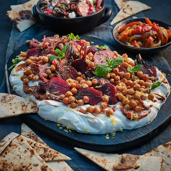 Plate of roasted beet salad with chickpeas, yogurt, herbs, and pita bread wedges on a dark surface.