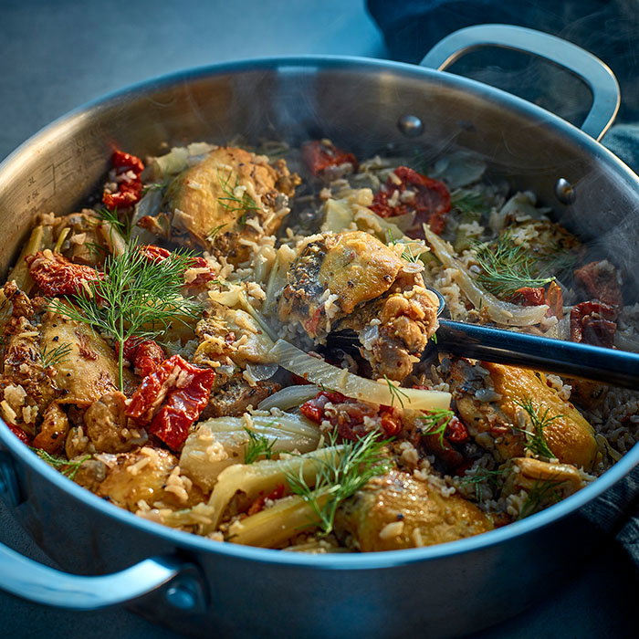 Steaming pot of cooked chicken with rice, dried tomatoes, fennel, and fresh dill garnish, with a spoon lifting a piece of chicken.