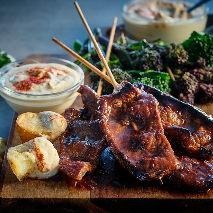 Grilled meat skewers with creamy dipping sauce, roasted potatoes, and roasted greens on a wooden serving board.