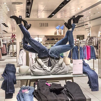 Creative retail display featuring mannequin legs in jeans positioned upside down on a white display table, surrounded by folded denim clothing in a contemporary clothing store
