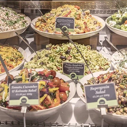 Deli counter display case with multiple prepared salads and dishes in round serving bowls, each with black price tags and labels