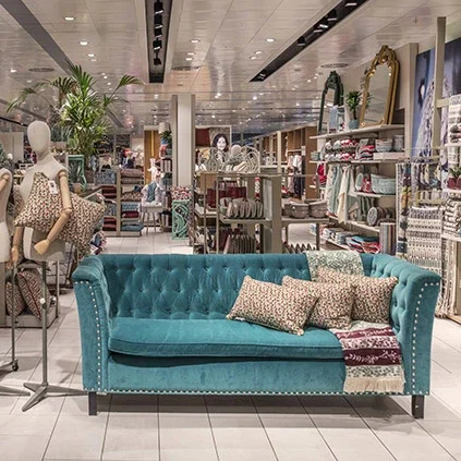 A teal velvet tufted sofa with decorative pillows displayed in a bright modern retail store with clothing racks and mannequins in the background