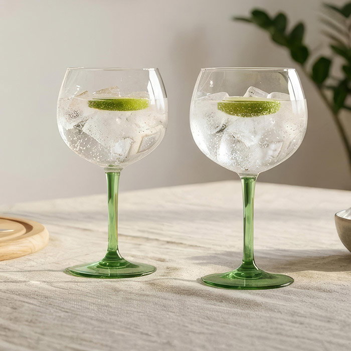 Two green-stemmed goblets filled with a fizzy drink, ice cubes, and lime slices sit on a textured table, with a soft-lit, cozy background.