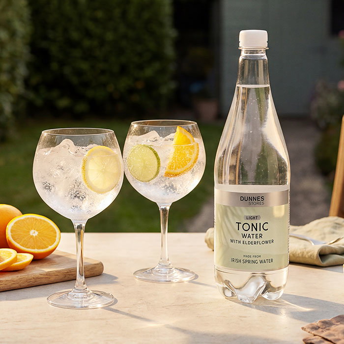 Two glasses of of clear water with ice and citrus slices beside a bottle of elderflower tonic water on an outdoor table.