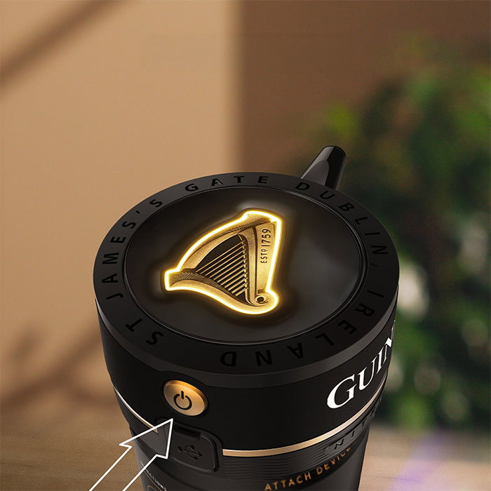 Close-up of a Guinness Nitrosurge device with illuminated harp logo and 'St James&rsquo;s Gate Dublin Ireland' text on top, with arrow pointing to the power button and charging port