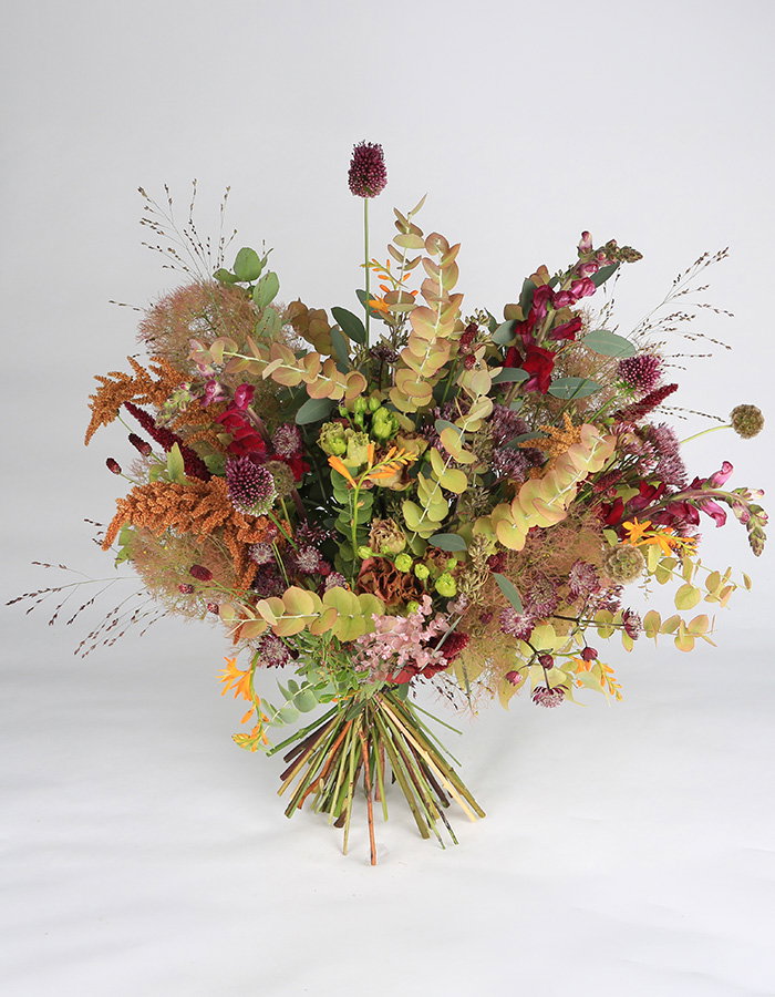 Lush, rustic wildflower bouquet with eucalyptus, berries, grasses, and dried blooms arranged in a loose fan, evoking warmth and abundance.