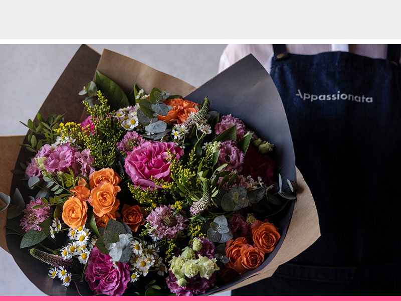 Colourful flower arrangement wrapped in brown paper featuring pink roses, orange roses, white daisies, and purple flowers, held by an Appassionata employee wearing a branded apron