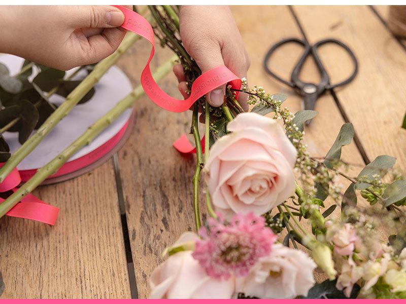 Hands arranging pink roses and small flowers with coral pink ribbon on a rustic wooden surface, with floral scissors visible in the background