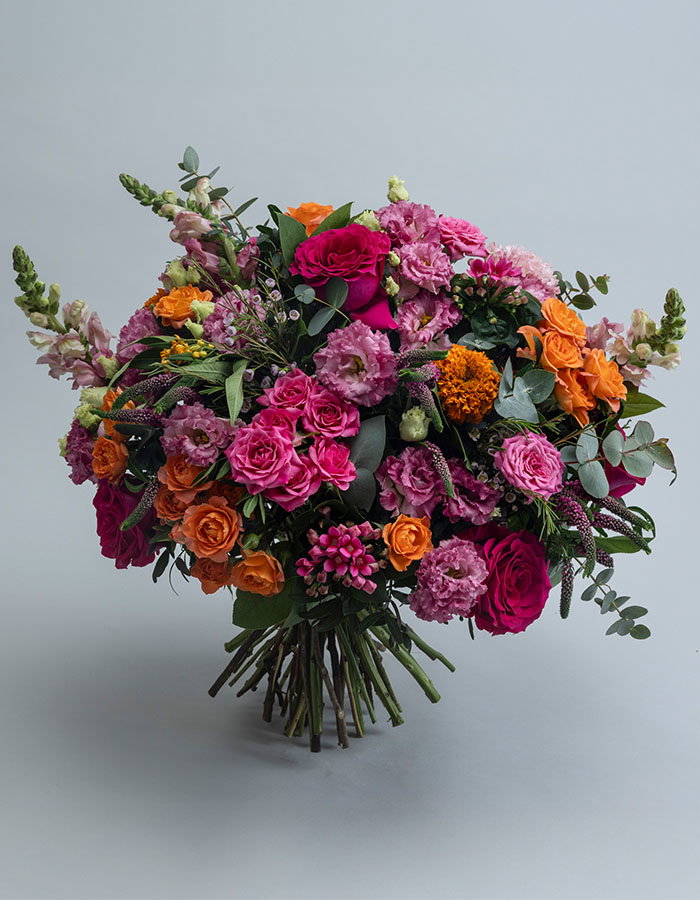 Vibrant mixed flower bouquet featuring deep pink roses, orange roses, purple carnations, and greenery against a light grey background