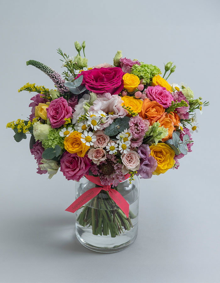 Mixed flower bouquet in a clear glass vase with pink ribbon, containing pink roses, yellow roses, white daisies, green hydrangeas, and various colourful blooms