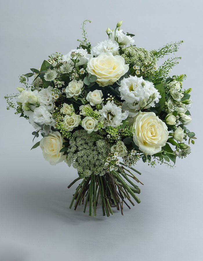 Elegant white and cream flower arrangement with white roses, white carnations, green foliage, and delicate filler flowers against a grey background