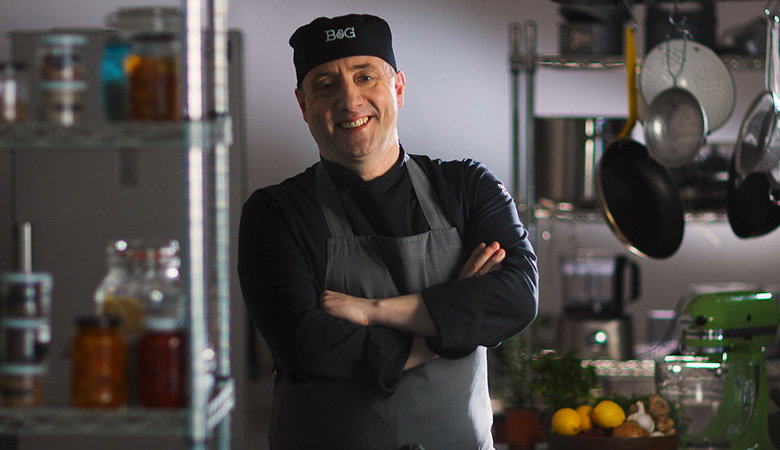 A smiling chef wearing a black chef hat and apron with arms crossed, standing confidently in a professional kitchen with pots, pans, and cooking equipment visible in the background