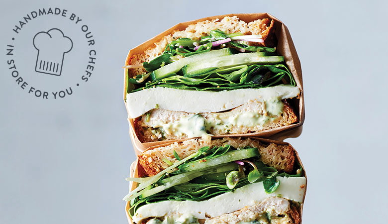 Two stacked gourmet sandwiches cut in half showing layers of fresh greens, cheese, and fillings on artisanal bread, with a circular logo reading 'HANDMADE BY OUR CHEFS - IN STORE FOR YOU'