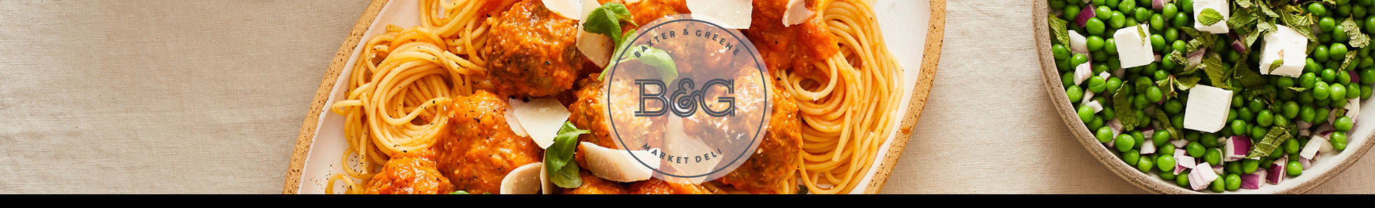 A plate of spaghetti with meatballs in tomato sauce, garnished with fresh basil and parmesan cheese shavings.  The Baxter & Greene logo is displayed on a white transparent circle in the centre of the image.