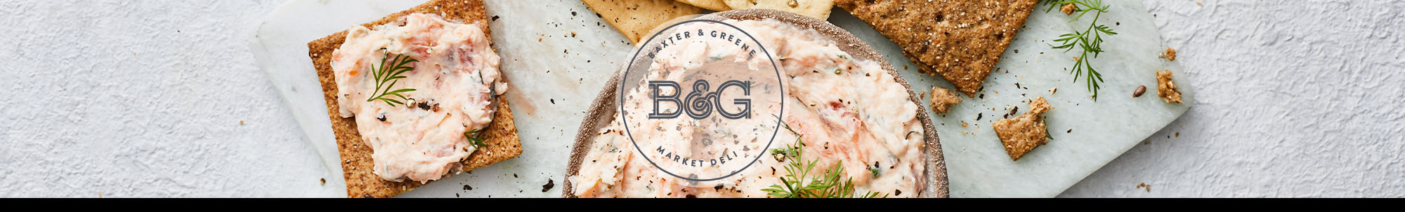 A bowl of creamy fish pâté garnished with fresh dill, served with assorted crackers and wine glasses.  The Baxter & Greene logo is displayed on a white transparent circle in the centre of the image.