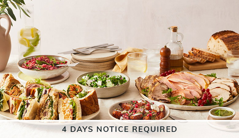 An elegant catering spread featuring sandwiches, salads, sliced meats, charcuterie, bread, and various accompaniments arranged on a white surface with the text '4 DAYS NOTICE REQUIRED' at the bottom