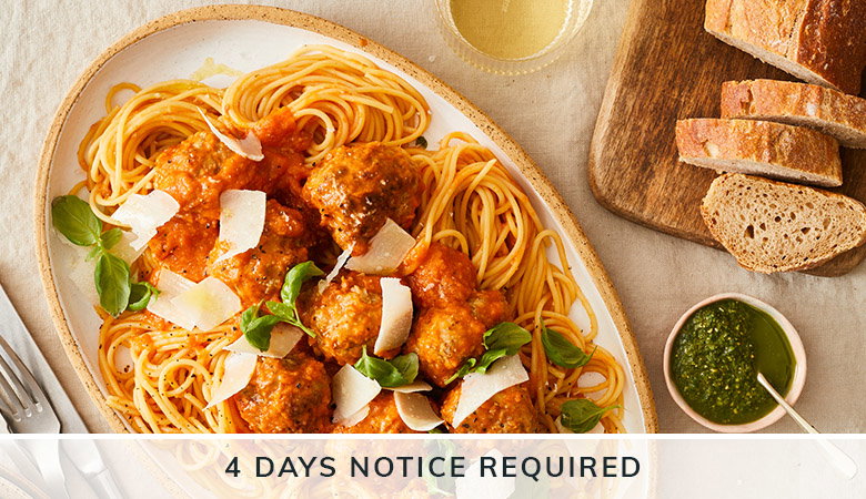 A plate of spaghetti with meatballs in tomato sauce, garnished with fresh basil and parmesan cheese shavings, served alongside bread and pesto, with '4 DAYS NOTICE REQUIRED' text overlay