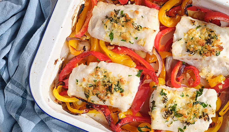 Baked white fish fillets topped with herbs and breadcrumbs, served over colorful roasted bell peppers in red and yellow, arranged in a white ceramic baking dish