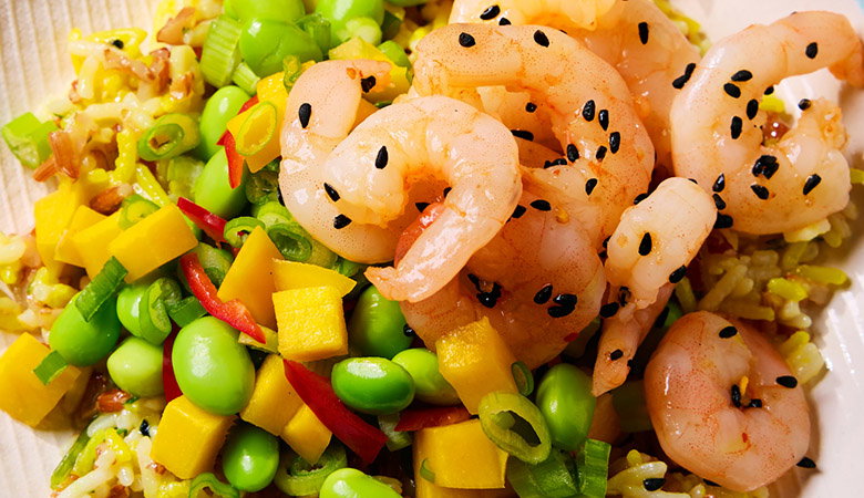 Colourful stir-fried rice dish with pink prawn, bright green peas, diced yellow bell peppers, and black sesame seeds, served in a white bowl