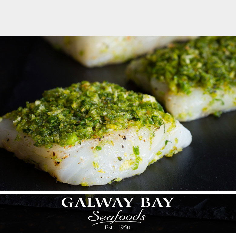 Fresh white fish fillets topped with vibrant green herb crust arranged on a dark slate surface, with additional herb-crusted fillets visible in the background. The Galway Bay Seafoods logo appears at the bottom in white serif typography with 'Est. 1950' beneath it
