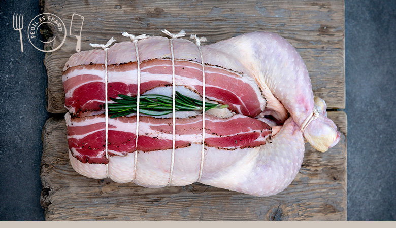 A whole chicken wrapped in bacon and tied with white butcher's twine, garnished with fresh rosemary, positioned on a rustic wooden cutting board.