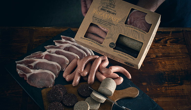 Artisanal charcuterie selection featuring sliced cured meats, various sausages, and black pudding arranged on a dark slate board, with a James Whelan branded gift box in the background on a rustic wooden surface.