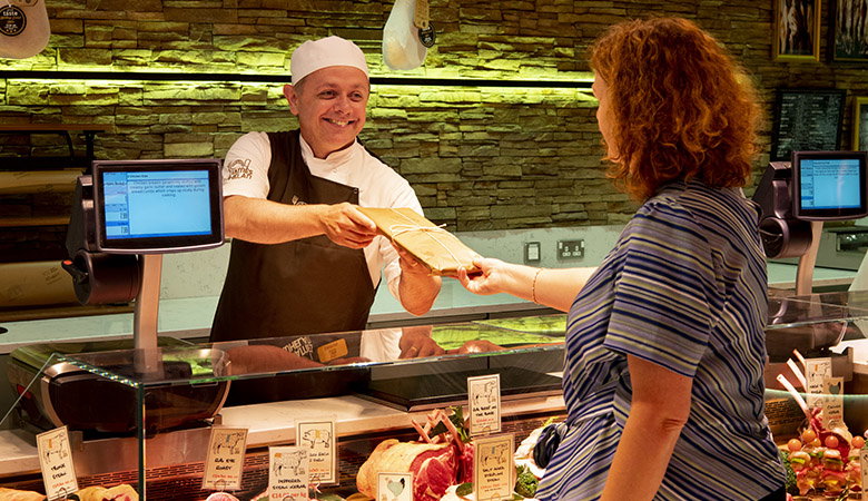 Friendly butcher in white uniform and cap serving a customer at a modern butcher shop counter, handing over wrapped meat with illuminated display cases and stone wall backdrop creating a warm, professional atmosphere.