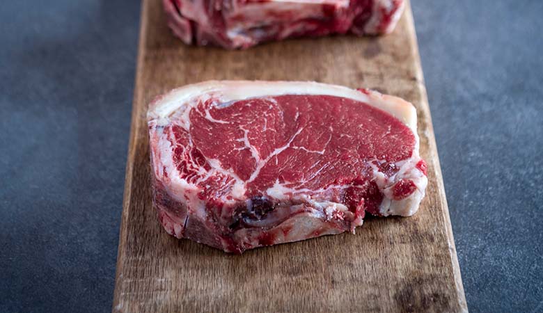 An elegant close-up shot of a raw, thick-cut ribeye steak with prominent white marbling, resting vertically on a rustic wooden board against a dark grey background.