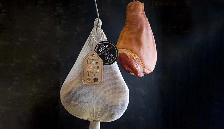 Whole cured ham and ham joint hanging against a dark background