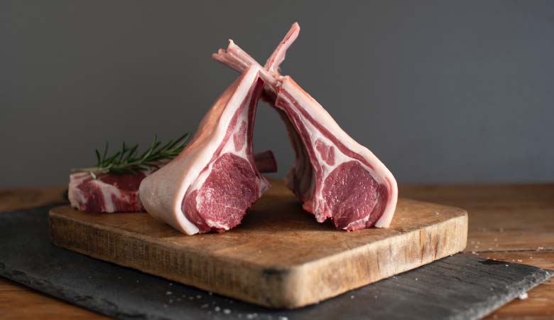Two raw, Frenched lamb racks leaning against each other on a rustic wooden cutting board with a sprig of rosemary in the background.