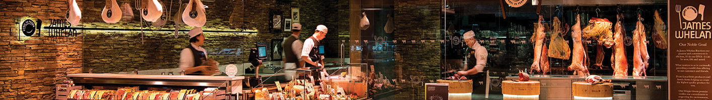 Panoramic view of a busy traditional butcher shop with multiple staff in white uniforms working at different stations, featuring hanging meats, display cases, and exposed brick walls creating an authentic artisanal atmosphere.