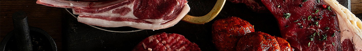 Gourmet meat platter featuring lamb chops, beef steaks, chorizo sausages, and other premium cuts artfully arranged on a dark wooden board with fresh herbs as garnish, showcasing variety and quality.