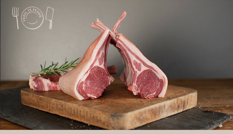 Two lamb chops with long frenched bones crossed together on a wooden cutting board, garnished with fresh rosemary, alongside additional chops.