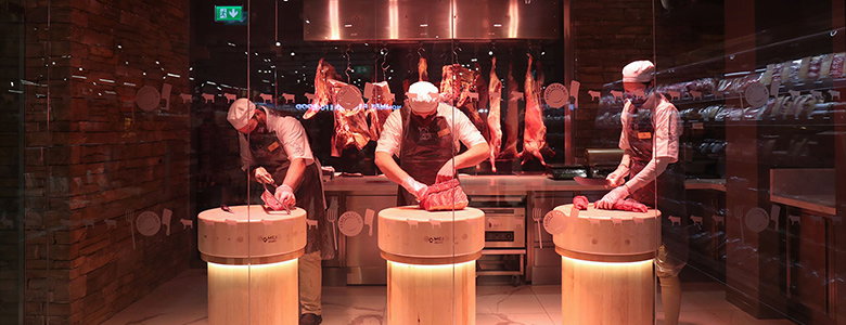 Professional butchers in white uniforms and caps working behind illuminated circular cutting stations in a modern butcher shop with warm lighting.