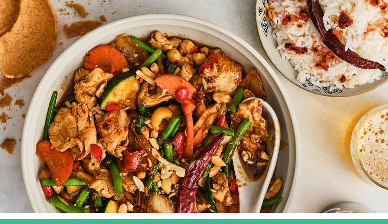 A colourful stir-fry dish in a white bowl featuring mixed vegetables, nuts, and protein with green beans, red peppers, and cashews, served alongside white rice and traditional ceramic dishes.