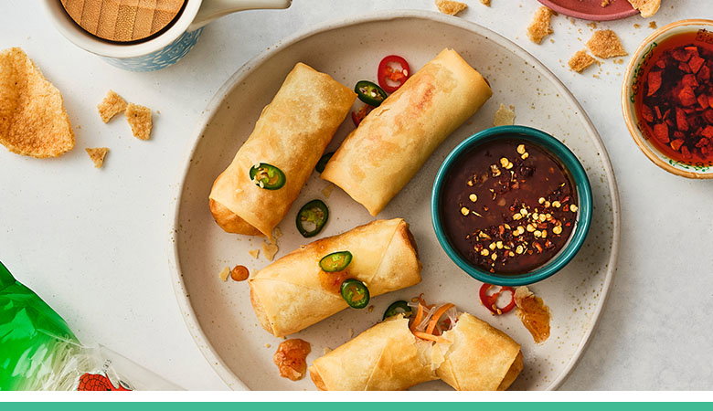 Four golden spring rolls arranged on a speckled ceramic plate, garnished with sliced jalapeños and cherry tomatoes, accompanied by a small bowl of dark dipping sauce with chilli flakes.