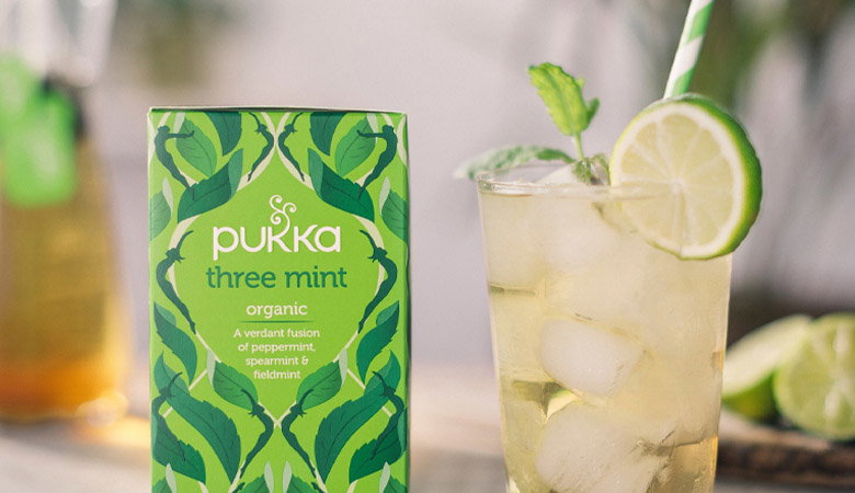 A glass of iced herbal tea with lime garnish and mint leaves next to a Pukka Three Mint Organic tea box featuring green leaf patterns on the packaging.