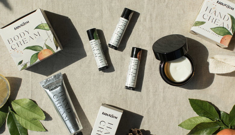 An artfully arranged flat lay of Nourish skincare products including tubes, jars, and boxes with green leaf branding, scattered on a neutral linen background with natural plant elements.