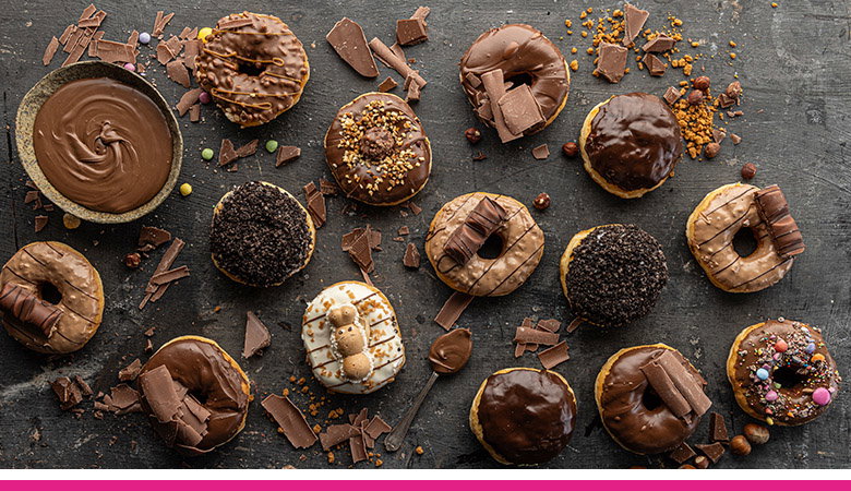 Overhead shot of various chocolate-covered donuts arranged on a dark surface, with chocolate shavings, a bowl of chocolate sauce, and a spoon. The donuts have different toppings, including nuts, sprinkles, and chocolate pieces.