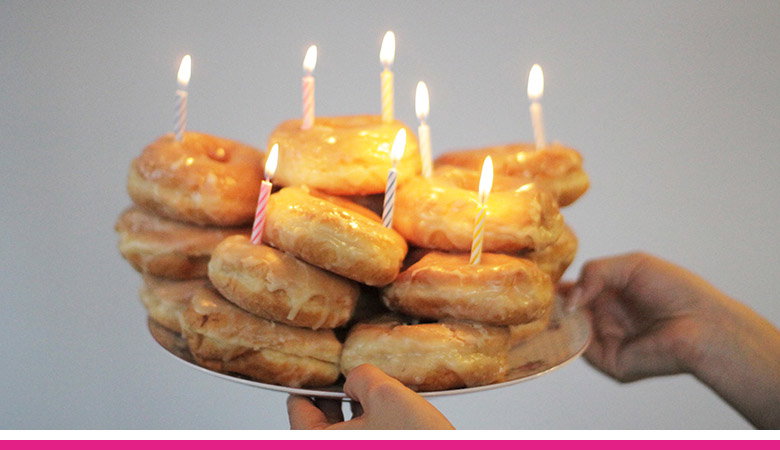 A stack of glazed donuts with lit candles on top, held by a person's hand. The donuts are arranged on a plate, and the background is a plain white wall.