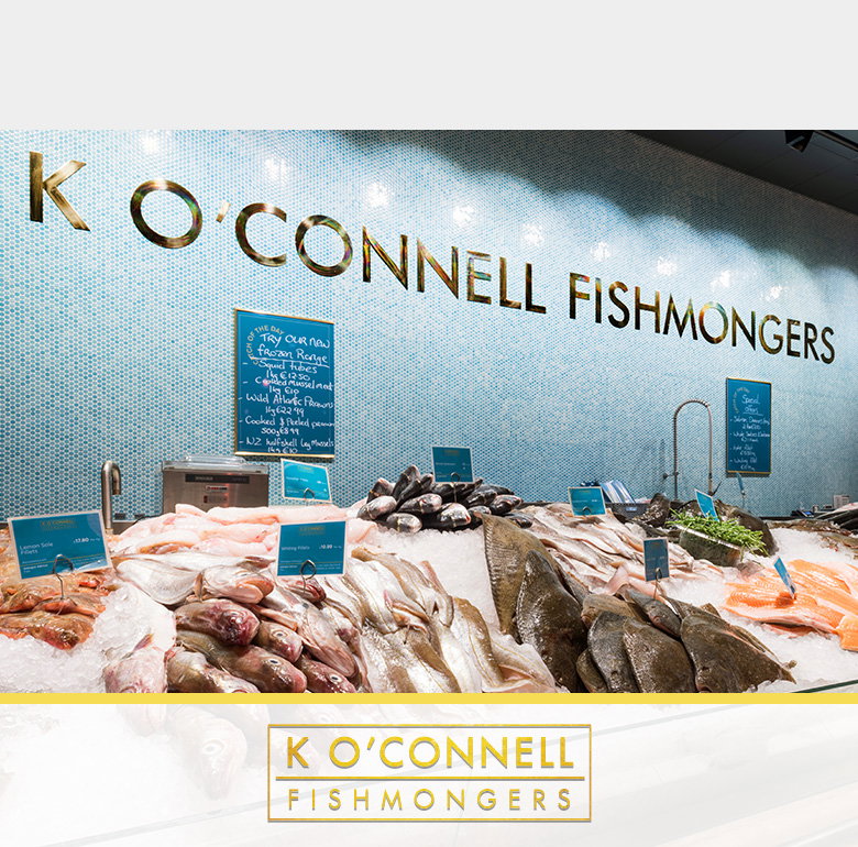 Interior view of K O'Connell Fishmongers with a curved glass display counter showcasing fresh fish and seafood on ice, with blue price tags throughout. The shop features white subway tiles and blue mosaic accent walls, with the business name prominently displayed above. Staff can be seen working behind the counter.