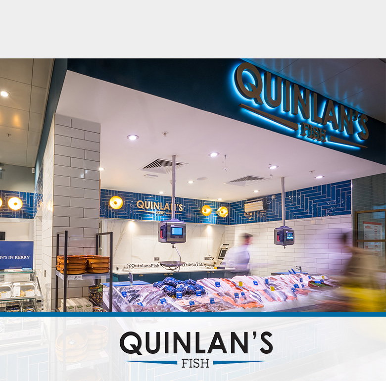 Modern Quinlan's Fish shop interior with contemporary design featuring white subway tiles, blue geometric wall patterns, and LED lighting. The curved seafood display counter shows fresh fish on ice with digital price displays, and motion blur suggests active customer service. The Quinlan's Fish logo appears at the bottom.