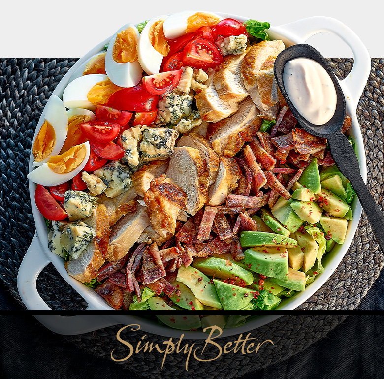 A gourmet salad bowl containing mixed greens, avocado slices, cherry tomatoes, hard-boiled eggs, blue cheese crumbles, bacon bits, and mushrooms, served with a small pitcher of dressing, promoting Simply Better's award-winning premium food collection exclusive to Dunnes Stores