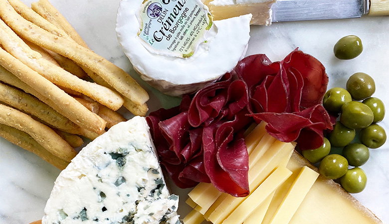 Elegant charcuterie board featuring artisanal cheeses including blue cheese and soft white cheese, thinly sliced cured meats, green olives, and golden breadsticks arranged on white parchment paper